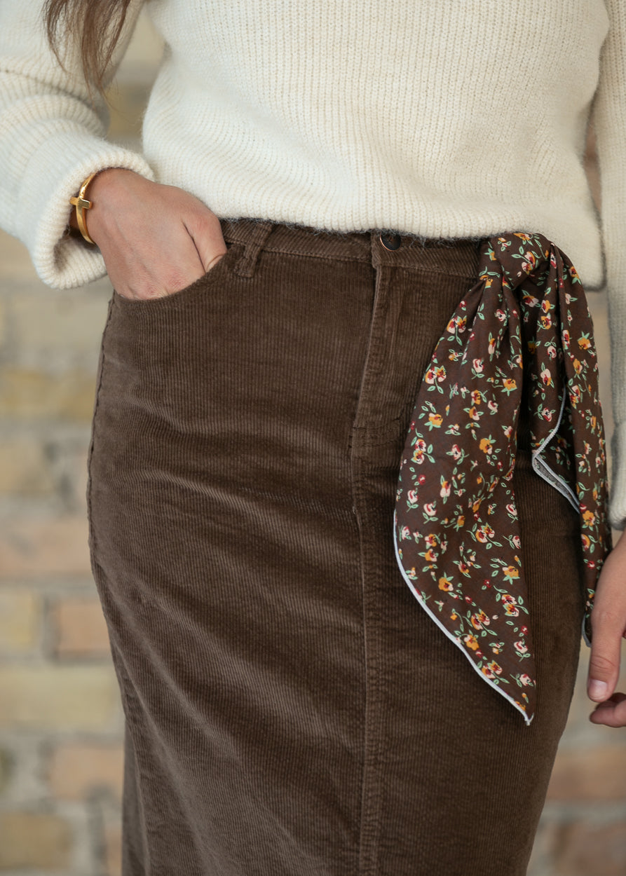 Close-up of brown corduroy maxi skirt with front pocket, belt loops, button closure, and floral scarf.