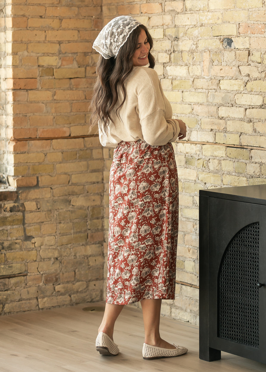 A woman wearing a cream knit top, a red and beige floral midi skirt, and a white lace headscarf stands in a rustic room with a brick wall and wooden floor.