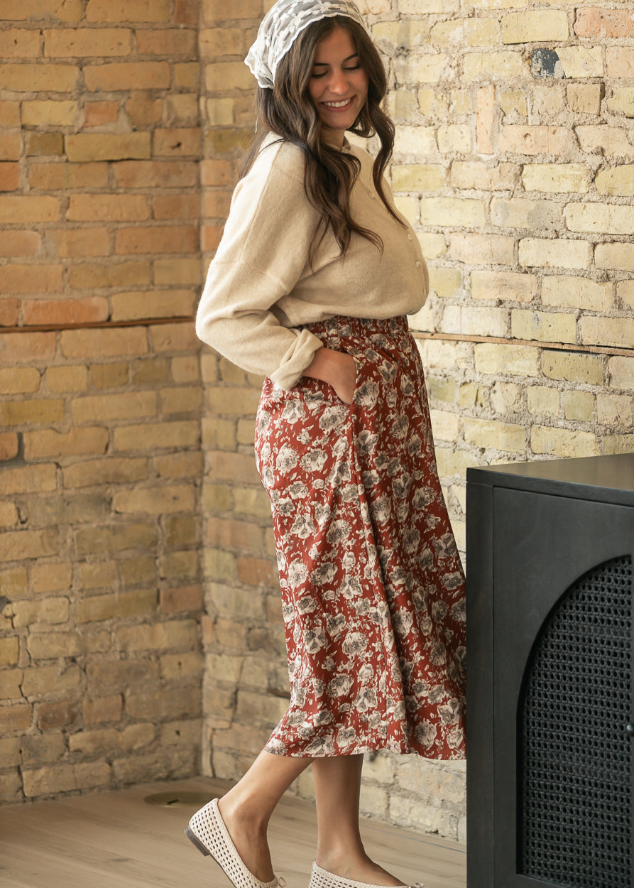 A woman wearing a beige knit sweater, a red and beige floral midi skirt with side pockets, a white patterned headscarf, and white woven flat shoes, posing in front of a rustic brick wall.