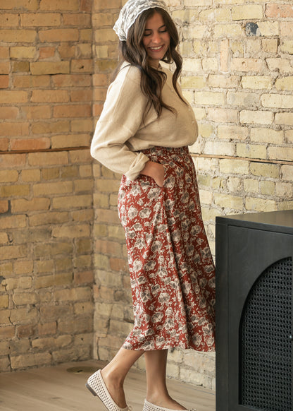 A woman wearing a beige knit sweater, a red and beige floral midi skirt with side pockets, a white patterned headscarf, and white woven flat shoes, posing in front of a rustic brick wall.