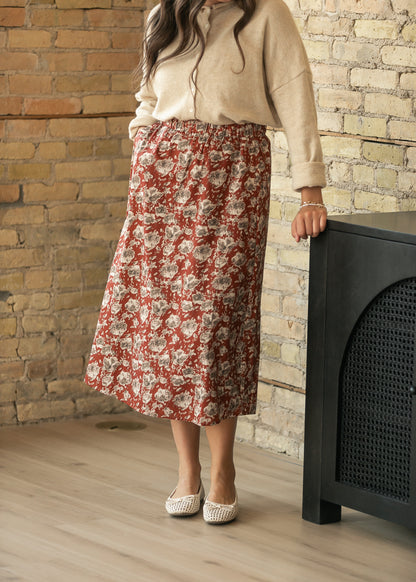 A woman wearing a beige long-sleeve top and a rust-red floral midi skirt, standing in a room with a brick wall and light wood floor next to a black cabinet.