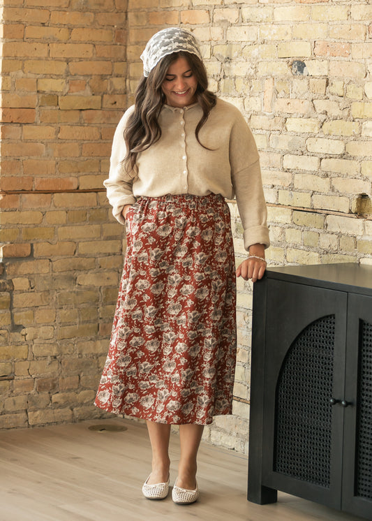 A woman stands in a rustic room with a brick wall, wearing a beige knit top, a red and beige floral midi skirt, a patterned headscarf, and white shoes.