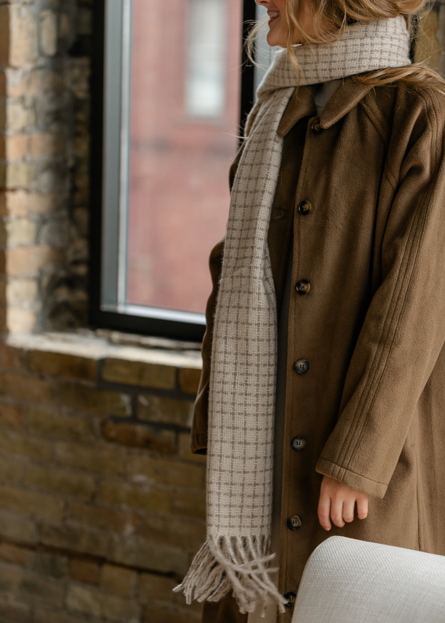 Neutral check scarf wrapped at the neck, showing its soft woven texture and long fringed ends.