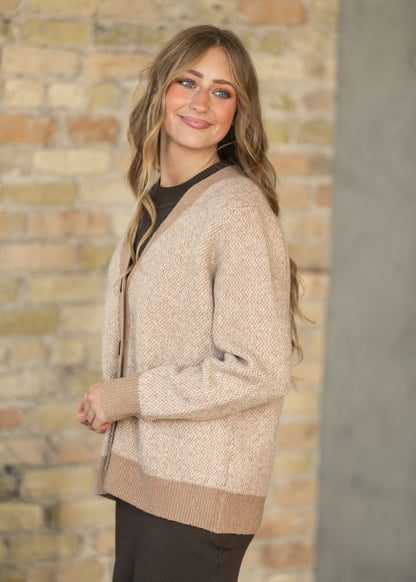 A woman wearing a beige textured knit cardigan over a black top, standing against a rustic brick wall background.