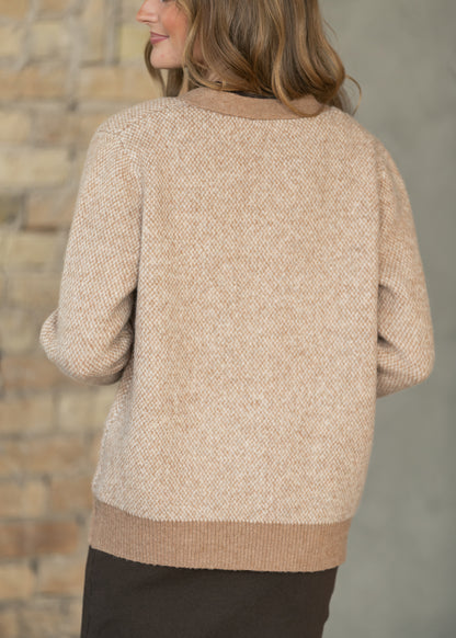 A woman shown from the back wearing a textured knit sweater with a beige and cream herringbone pattern and solid tan ribbed collar, hem, and cuffs, set against a blurred brick wall background.