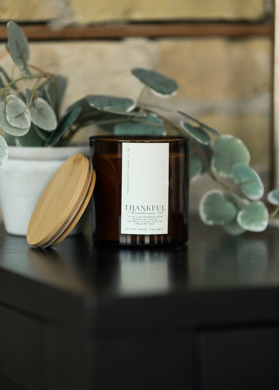 Amber Thankful soy candle with scripture-inspired label and natural wood lid displayed beside the jar.