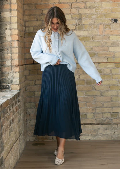 Deep navy pleated midi skirt with a loose flowing silhouette, paired with a soft blue cardigan and flats.