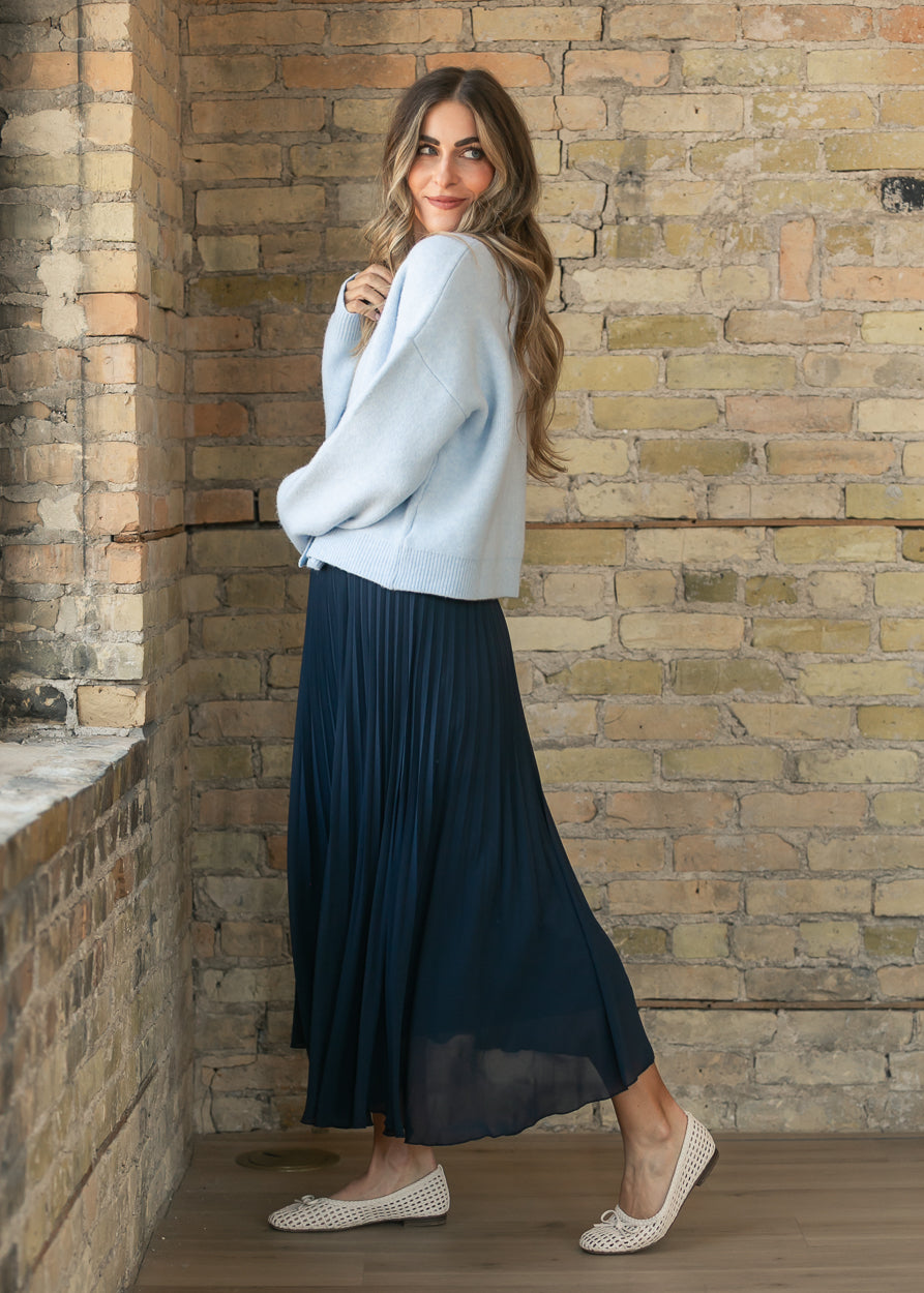 Navy pleated midi skirt shown from the side with a light blue cardigan and neutral woven flats.