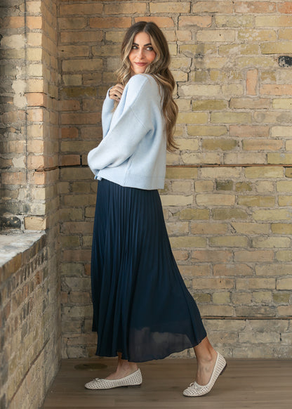 Navy pleated midi skirt shown from the side with a light blue cardigan and neutral woven flats.