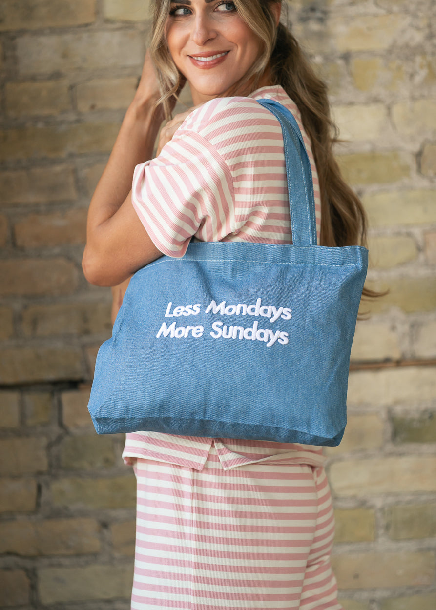Medium wash denim shoulder bag with white "Less Mondays More Sundays" embroidery.