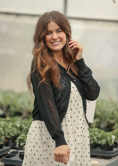 Black cropped cardigan layered over a cream polka dot maxi dress with ballet flats