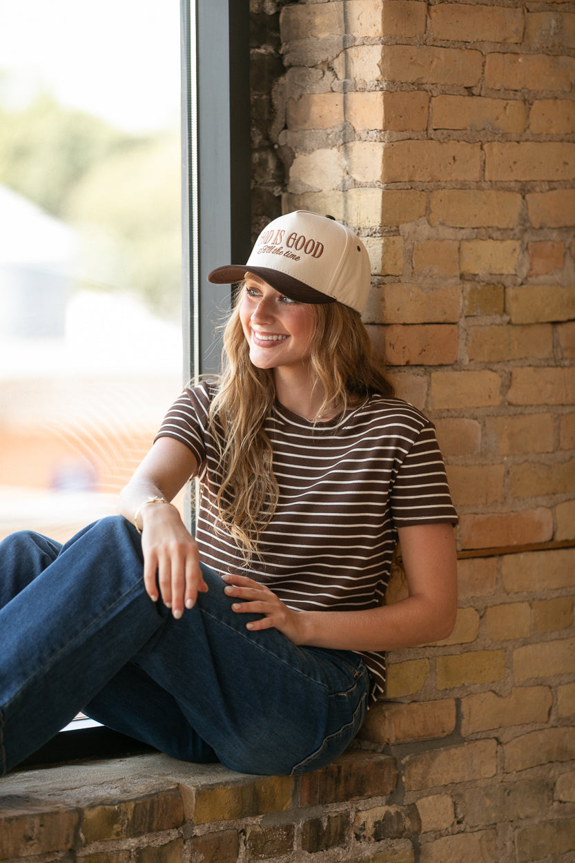 Mahogany brown striped short sleeve top styled with blue jeans while seated by a brick window.