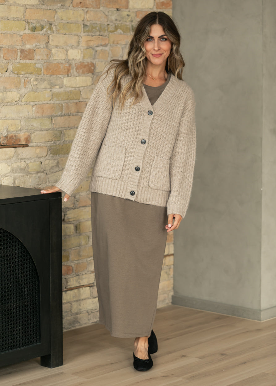 A woman models a beige ribbed cardigan over a taupe long-sleeve top and matching taupe maxi skirt, standing in a room with a brick and concrete wall.