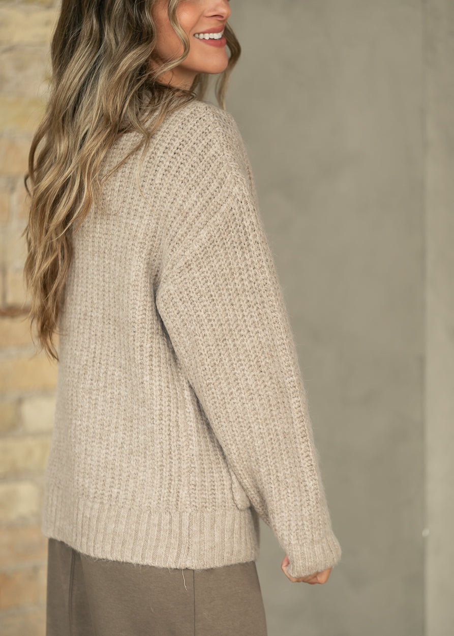 A woman wearing a chunky knit beige sweater, shown from a side-back angle against a neutral gray wall.