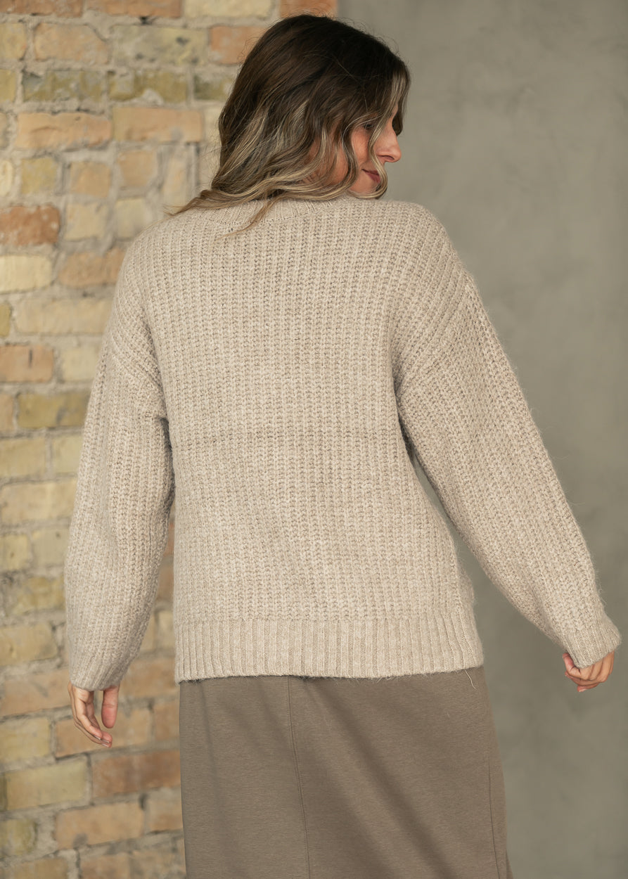 A woman shown from the back wearing a chunky knit beige sweater and a taupe skirt, standing against a split background of a brick wall and a gray wall.