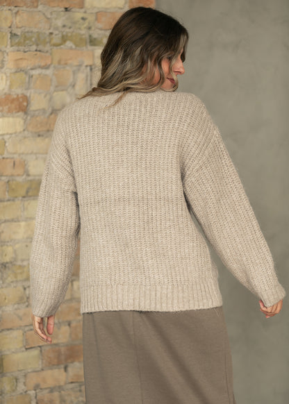 A woman shown from the back wearing a chunky knit beige sweater and a taupe skirt, standing against a split background of a brick wall and a gray wall.