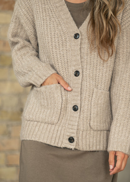 A close-up of a person wearing a chunky knit cardigan in a warm taupe-beige color. The cardigan features a V-neck, dark buttons, and front patch pockets, layered over a matching taupe dress or sweater.