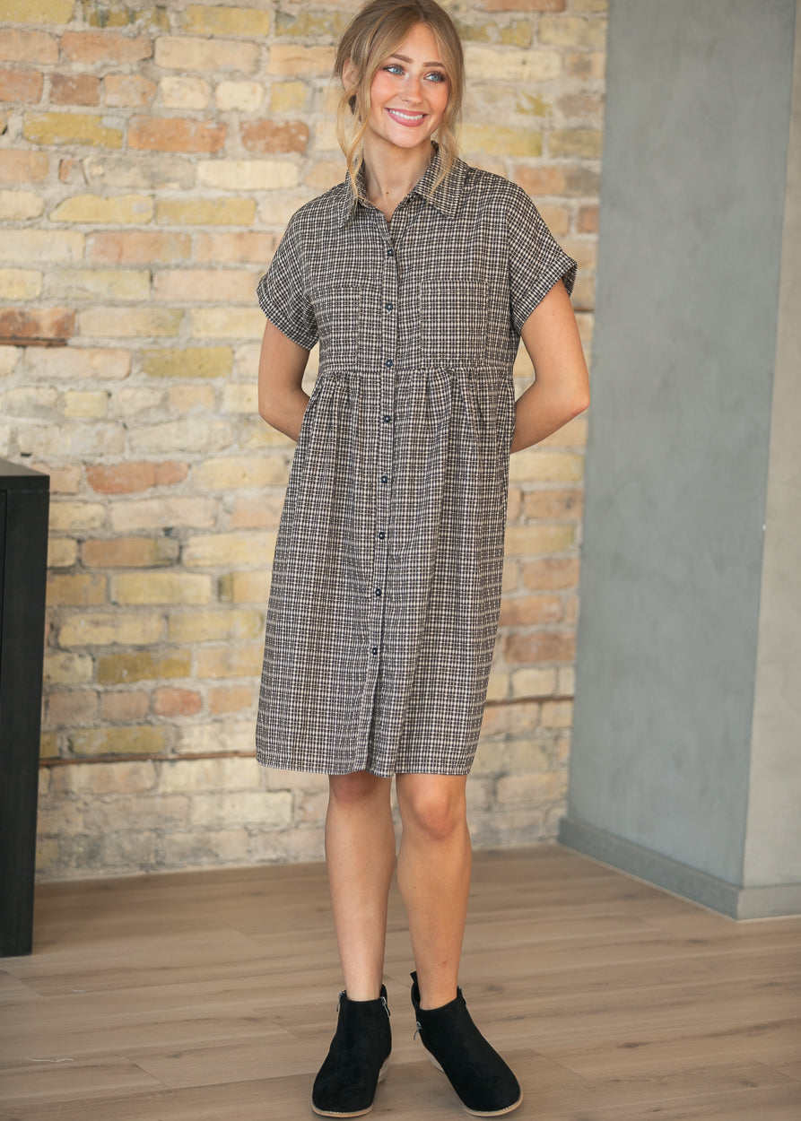 A woman wearing a short-sleeve button-front dress with a gray and white windowpane plaid pattern, standing in front of a brick wall.
