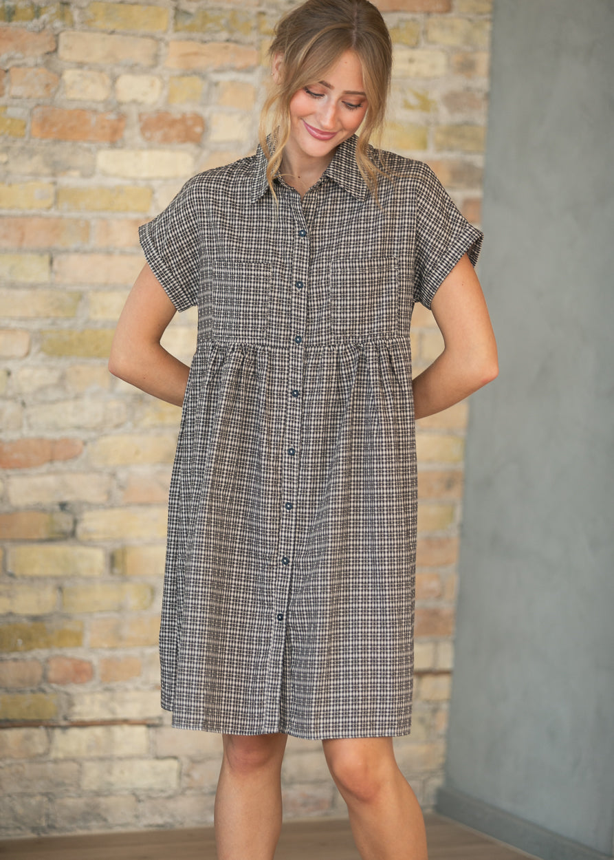 A woman wearing a short-sleeve button-front shirt dress with a gray and white windowpane plaid pattern, standing in front of a brick wall.
