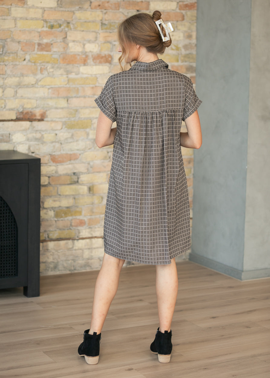 A woman viewed from behind wearing a short-sleeve collared dress with a brown and beige windowpane plaid pattern, paired with black ankle boots, standing in front of a brick wall.