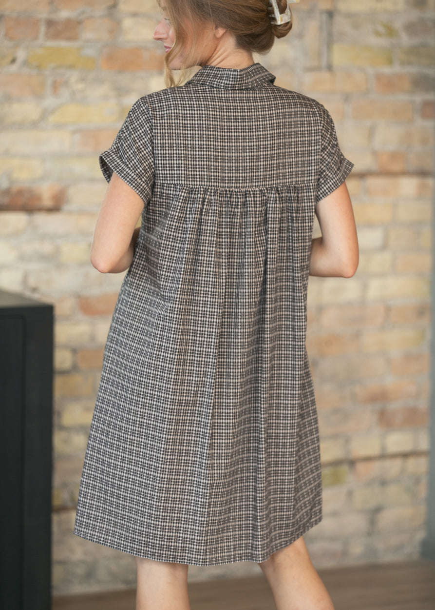 A woman wearing a short-sleeve plaid shirt dress, shown from the back. The dress has a brown, gray, and white checkered pattern and is photographed against a rustic brick wall background.