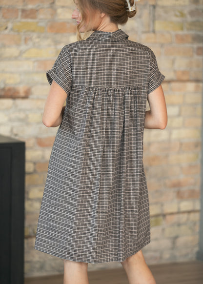 A woman wearing a short-sleeve plaid shirt dress, shown from the back. The dress has a brown, gray, and white checkered pattern and is photographed against a rustic brick wall background.
