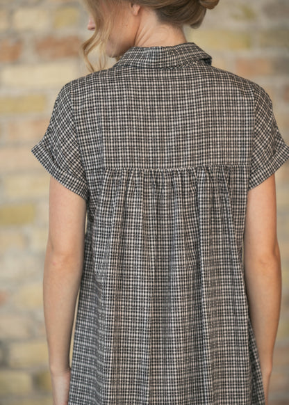 A woman shown from behind wearing a short-sleeve plaid shirt dress in brown, beige, and black tones, set against a blurred brick wall background.