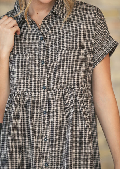 A close-up of a person wearing a short-sleeve button-front shirt dress with a small-scale black and white gingham pattern. The dress features a collar, two chest pockets, and a gathered waist seam.