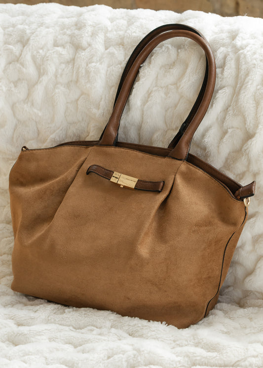 Brown faux suede tote with dark brown handles and gold-tone front accent, resting on a cream blanket.