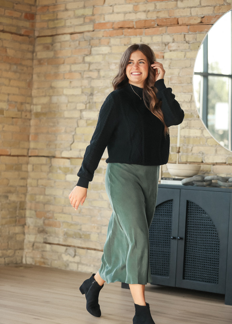 Model wearing an emerald faux suede midi skirt with a black sweater, walking view