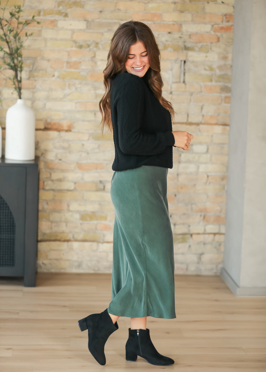 Full-length side view of emerald faux suede midi skirt with black sweater and ankle boots