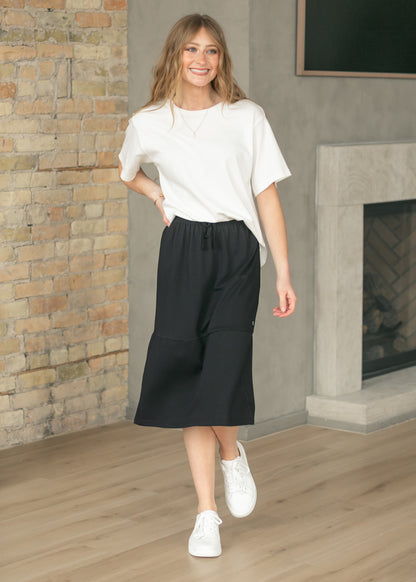Black midi sweat skirt styled with a white tee and sneakers beside a brick fireplace.