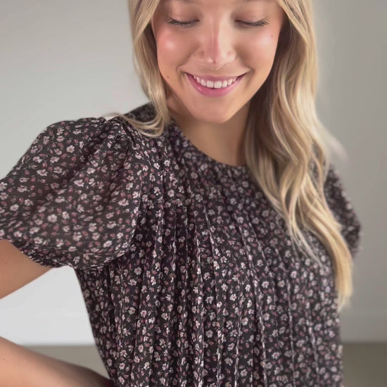 A black floral short sleeve top that is fully lined, with a round neckline, keyhole closure, and a pleated bodice.