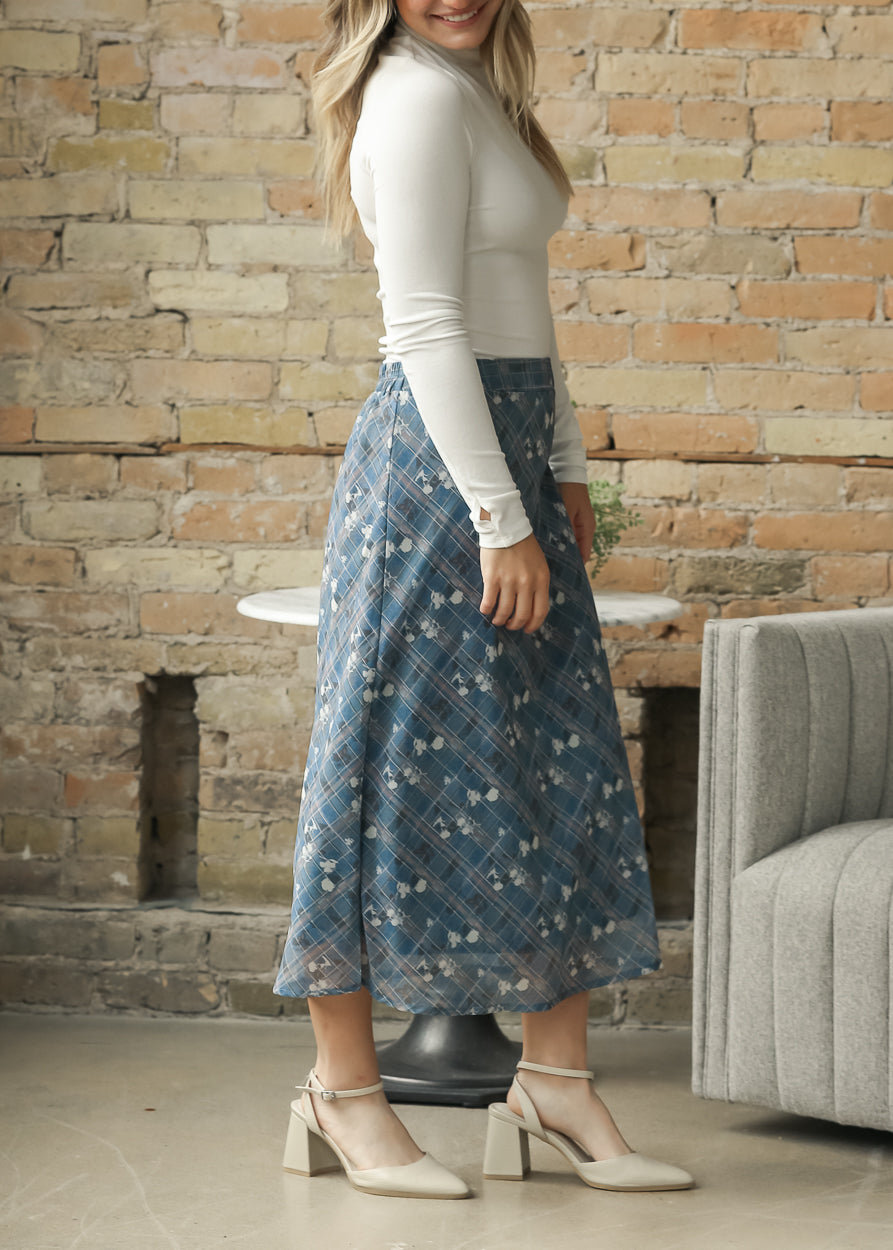 A woman wearing a white long-sleeve top and a blue floral midi skirt with white block-heel mules.