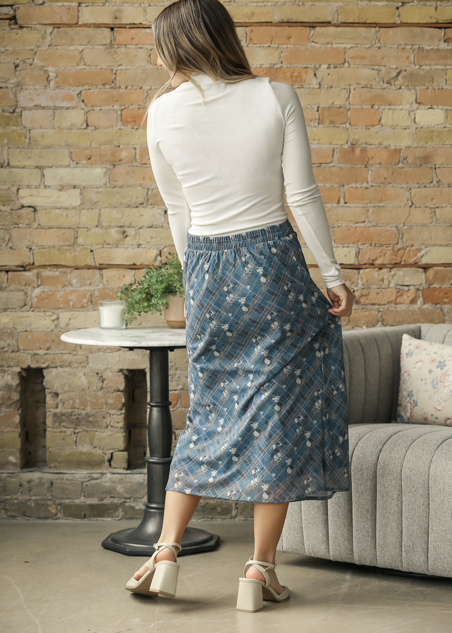 A woman stands with her back to the camera in a living room setting, wearing a white long-sleeve top and a blue floral midi skirt with a plaid waistband. She is wearing beige block-heel sandals