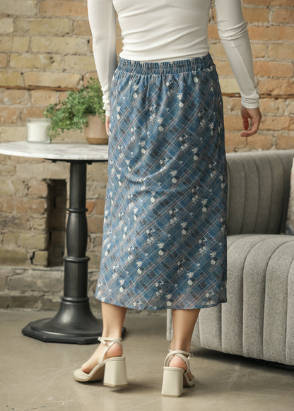 A person wearing a blue plaid midi skirt with a white floral pattern, paired with a white long-sleeve top and beige block-heel sandals.