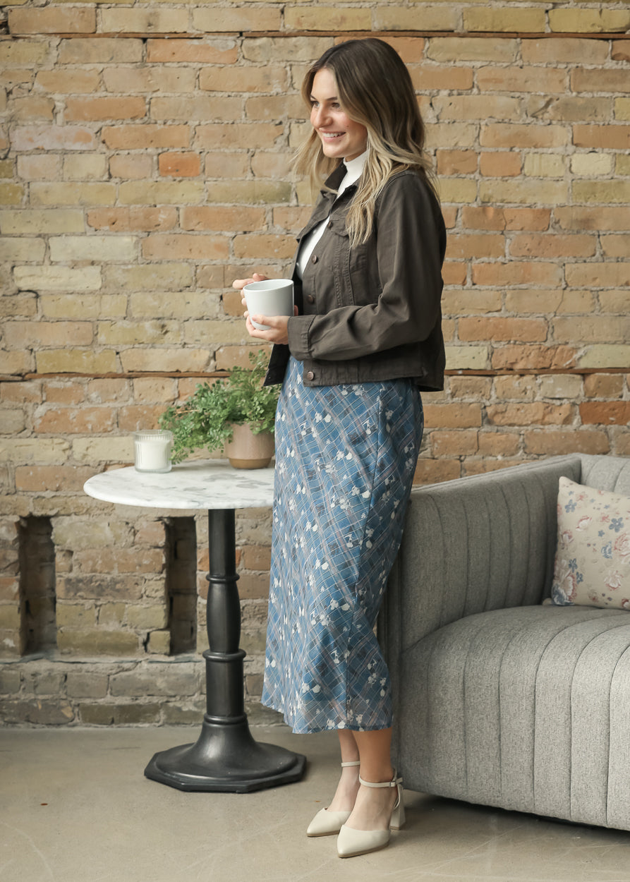 A woman stands in a cozy room with a brick wall background, wearing a dark brown cropped jacket, a white collared shirt, and a blue floral midi skirt. 