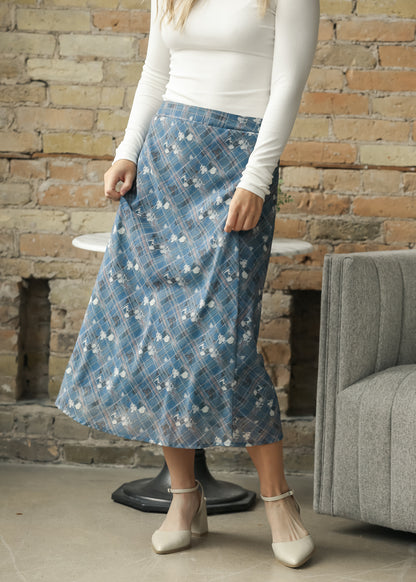 A woman wearing a blue floral midi skirt with a plaid waistband, paired with a white long-sleeve top and beige heels.