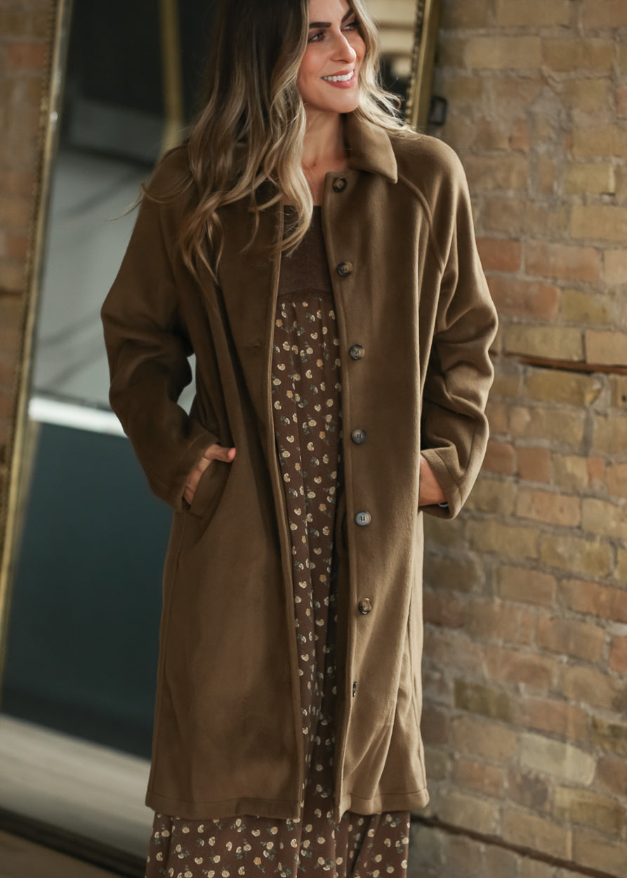 A woman wearing a brown mid-length button-front coat over a brown floral dress.
