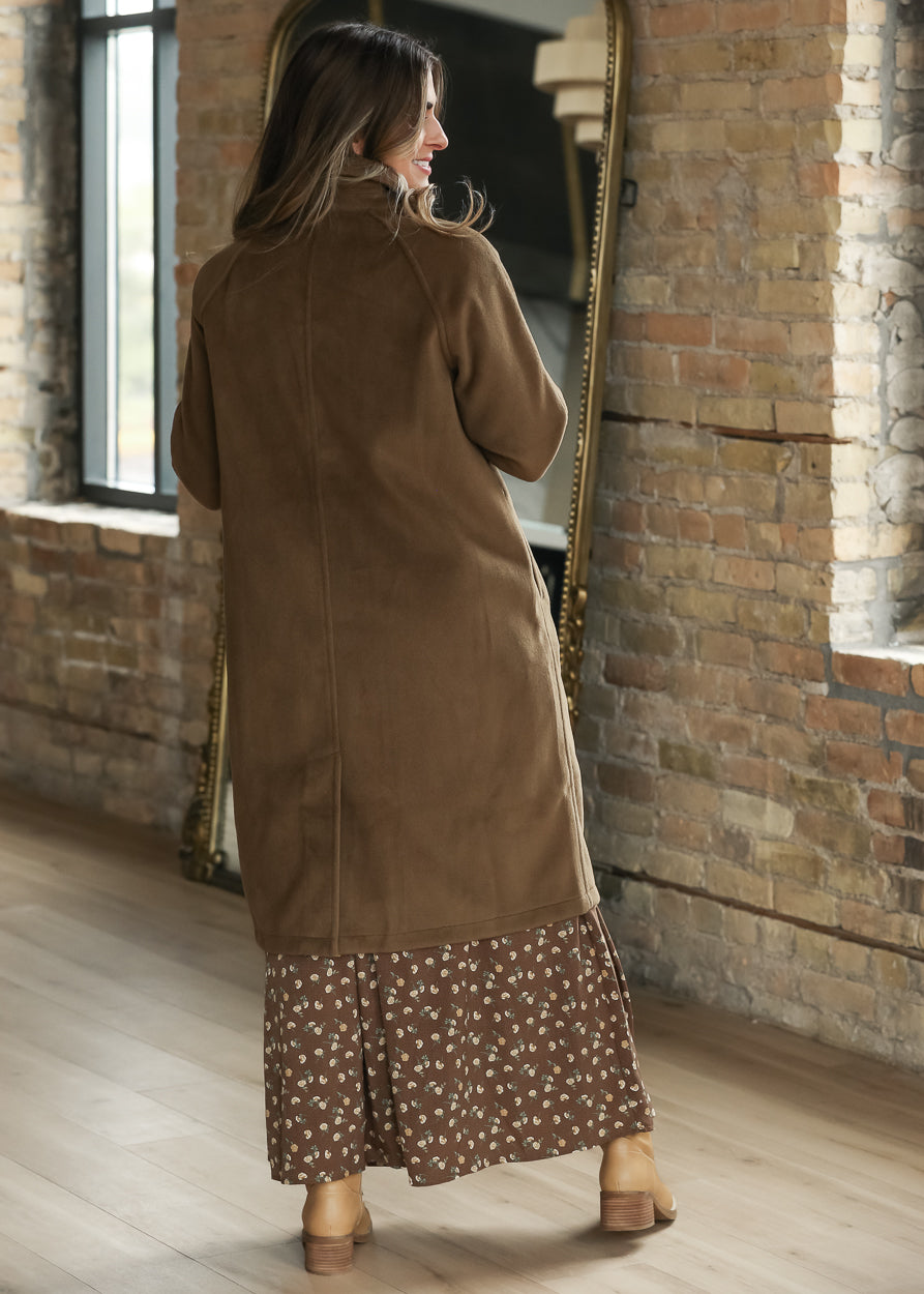 A woman stands with her back to the camera wearing a long brown suede-like cardigan over a brown floral maxi dress.