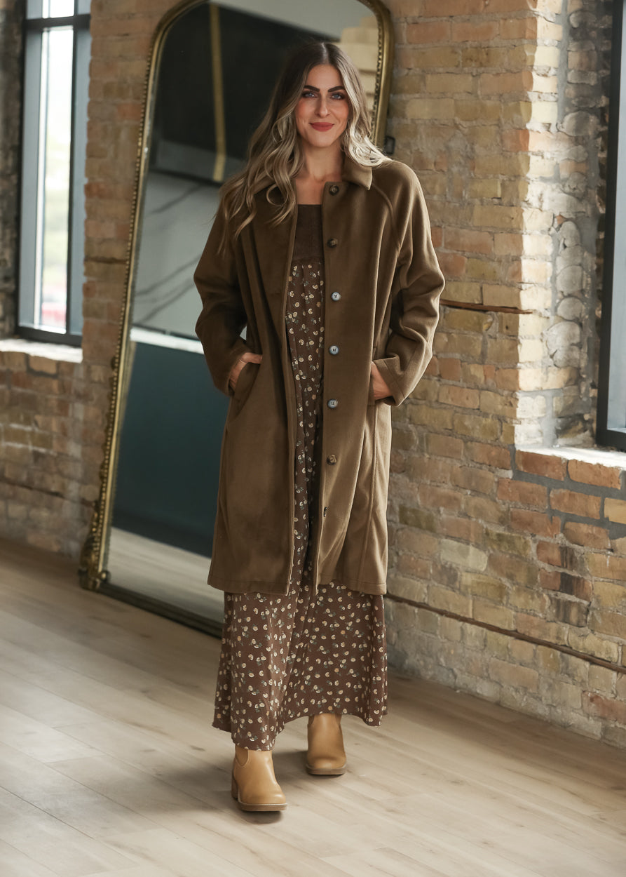 A woman wearing a long brown coat over a brown patterned dress, paired with tan ankle boots. 