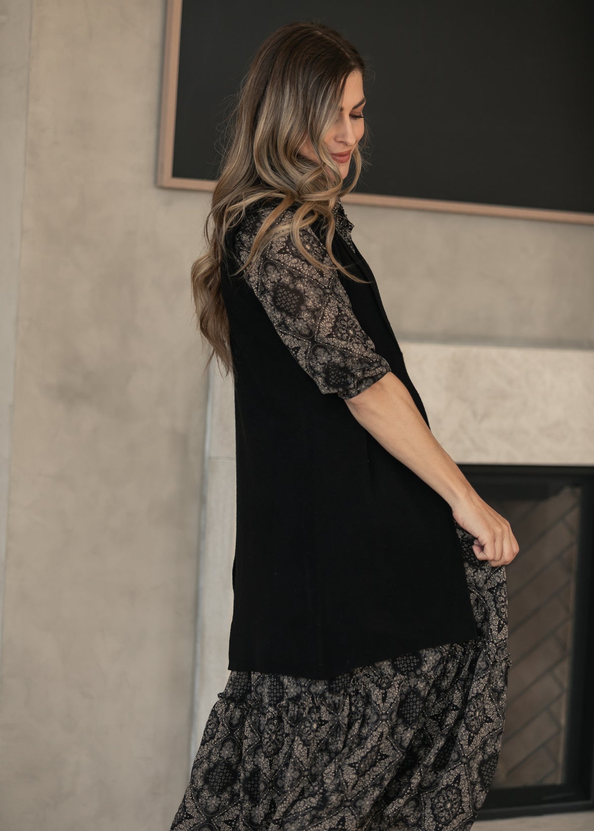 Side of black long cardigan showing its sleeveless cut and hem slit, layered over a paisley maxi dress.