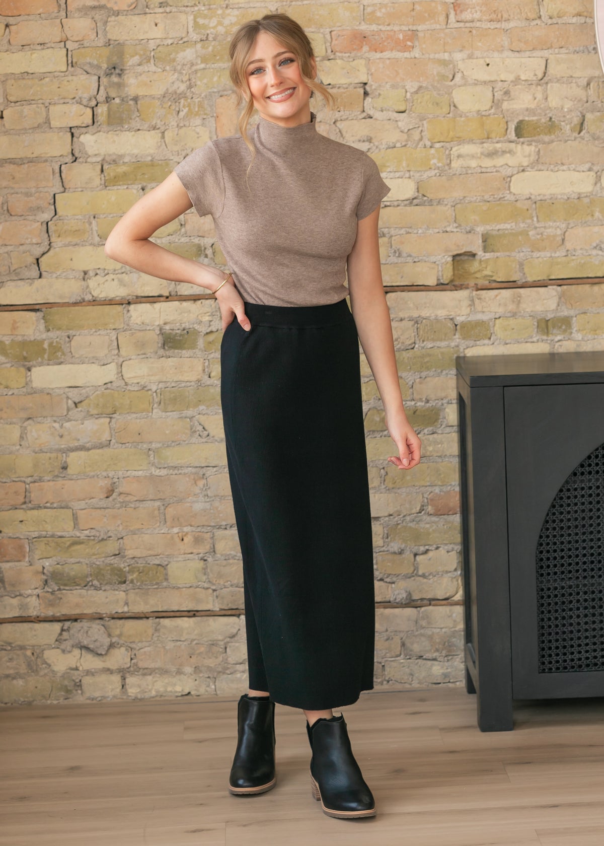 Woman wearing a brown top and black skirt standing against a brick wall.