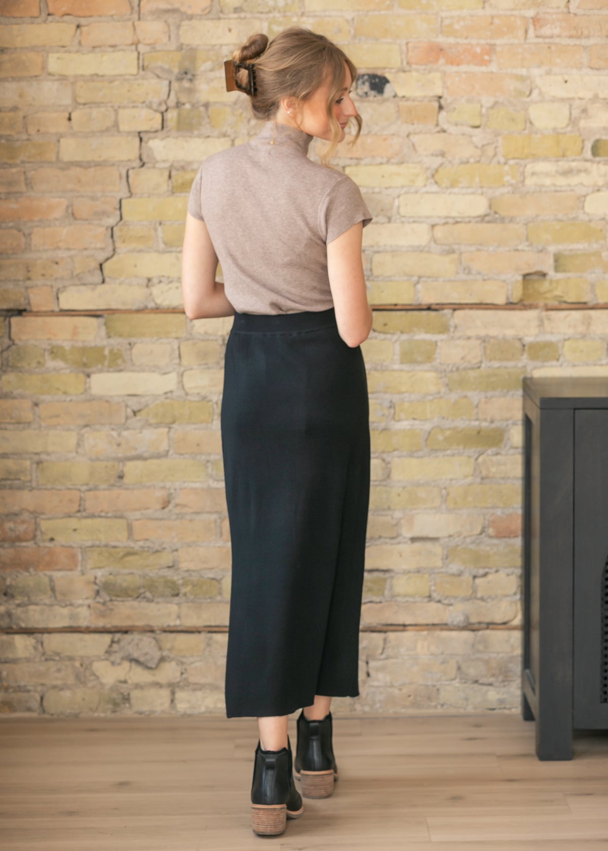 Back view of a woman wearing a beige top and black skirt against a stone wall.
