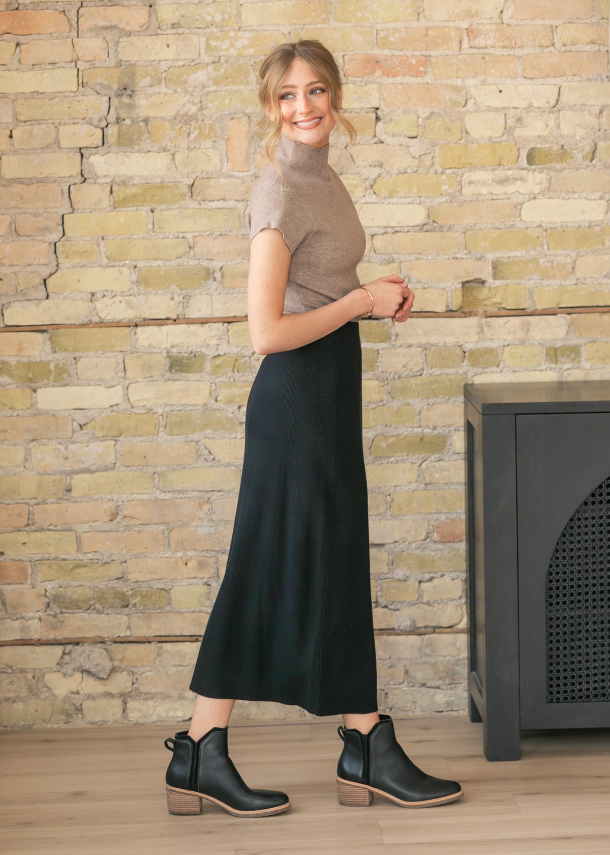 Side view of a woman in a beige top and black skirt standing against a brick wall.
