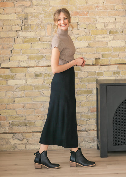 Side view of a woman in a beige top and black skirt standing against a brick wall.