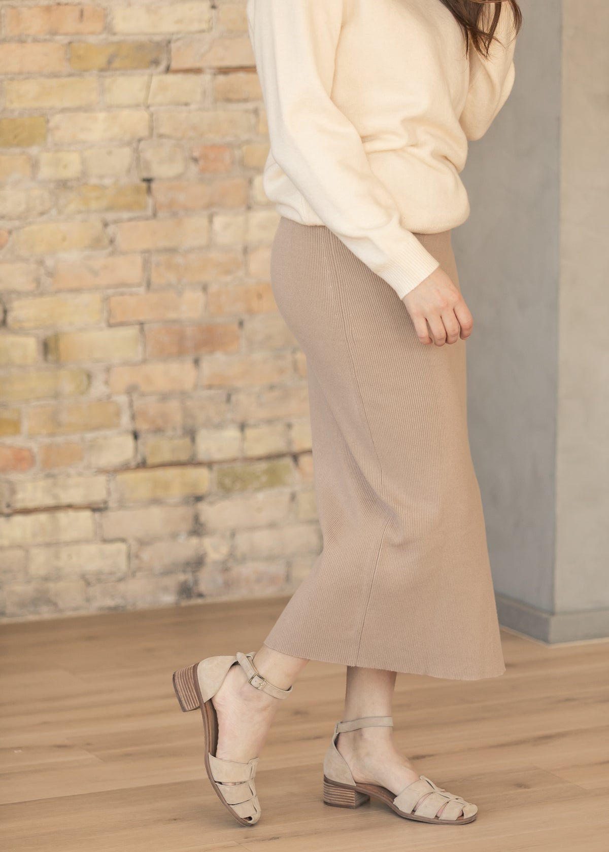 Side view of a beige skirt and white sweater against a brick wall.