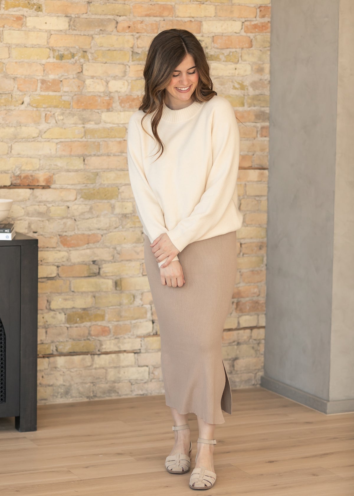 Woman wearing a cream sweater and beige skirt standing against a brick wall.