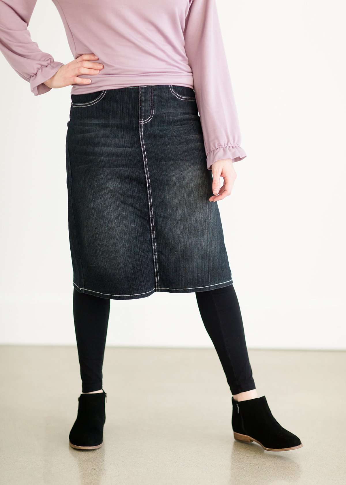 A black wash denim skirt with a ribbed knit elastic waist band. This skirt is paired with a purple sweatshirt, black leggings and black boots.