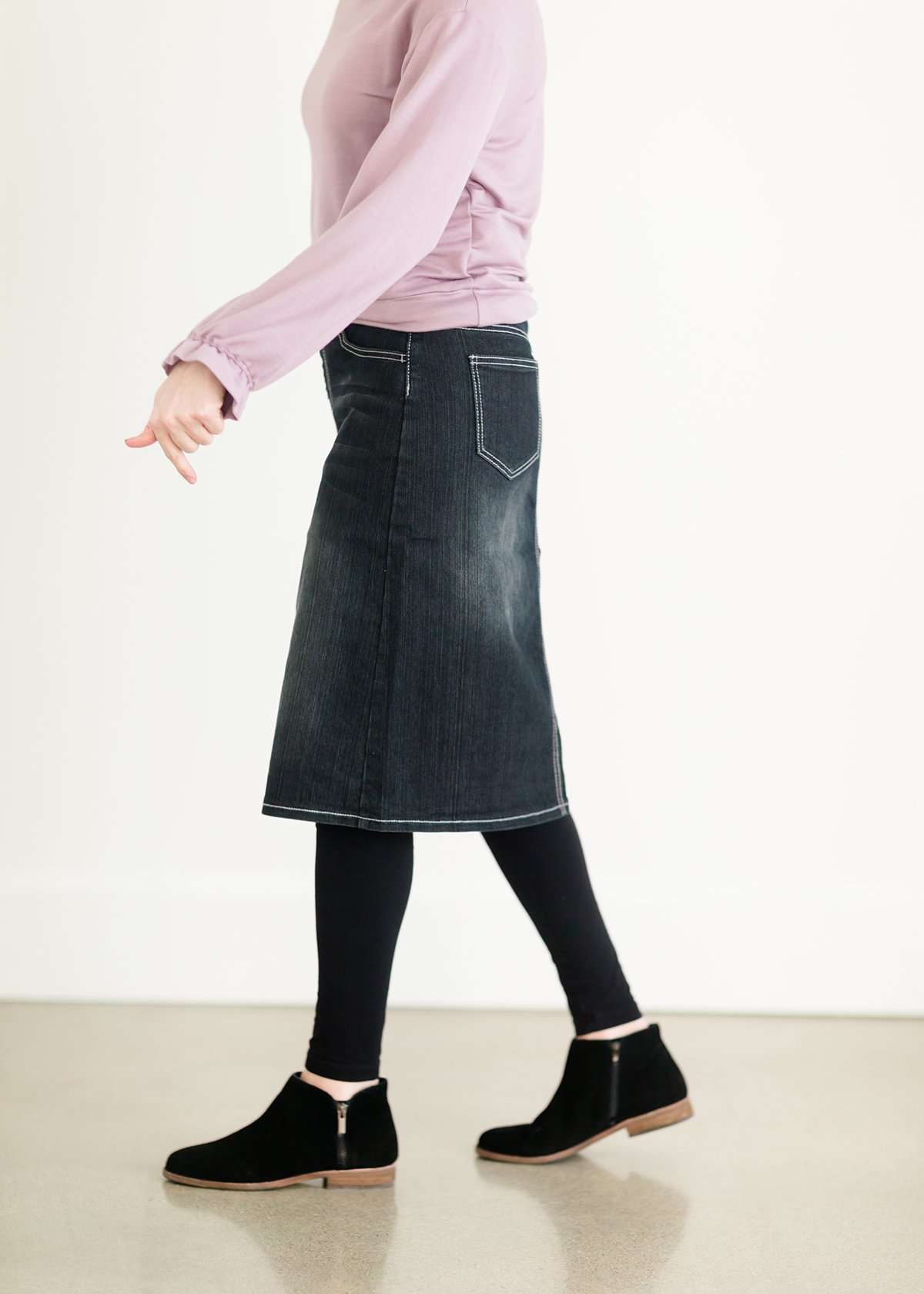 A black wash denim skirt with a ribbed knit elastic waist band. This skirt is paired with a purple sweatshirt, black leggings and black boots.